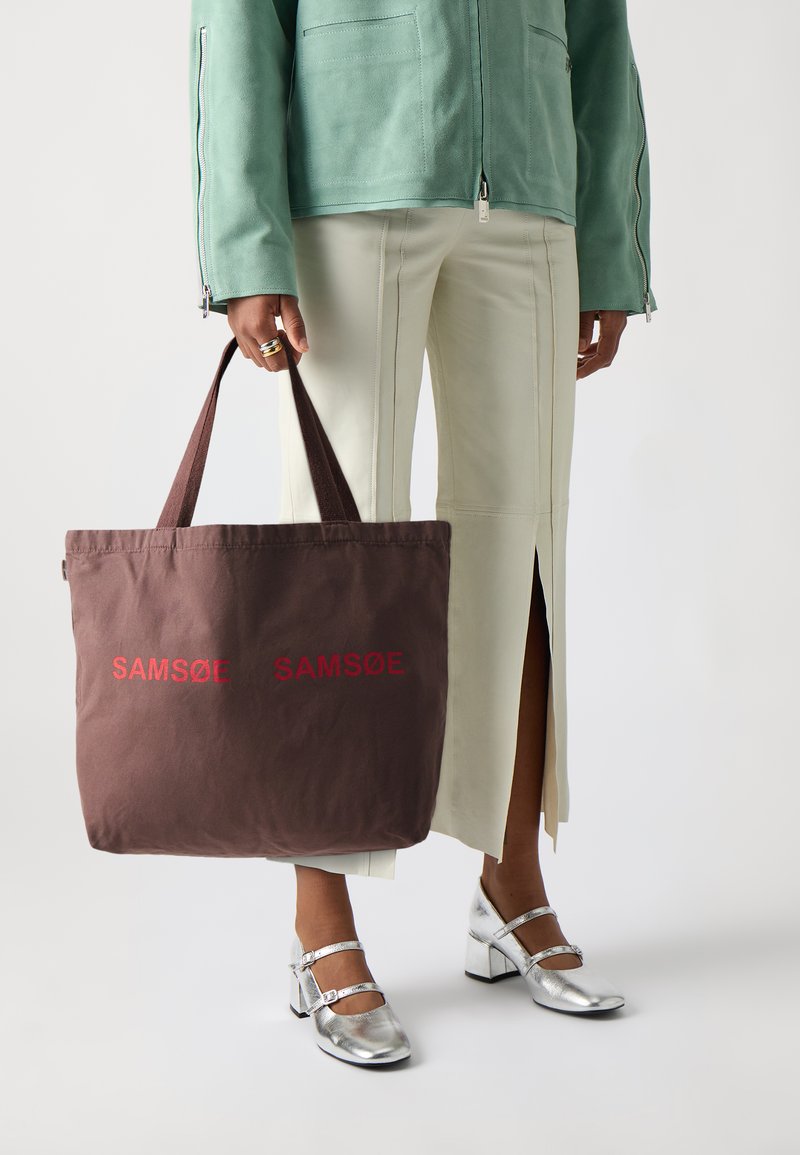 Bruine canvas draagtas met rode tekst "SAM SøE". Kenmerkt stevig materiaal, brede hengsels en een open bovenkant. Model draagt lichtgekleurde broek en zilveren schoenen.