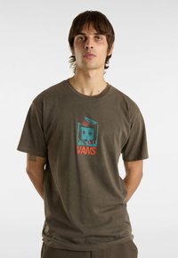 Olivově zelené bavlněné tričko s kulat�ým výstřihem, na kterém je barevný grafický potisk obličeje a text "VANS" uprostřed. Krátké rukávy.