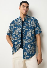 Camicia a maniche corte in tessuto blu scuro con motivi floreali beige, caratterizzata da una chiusura frontale con bottoni e orlo arrotondato, abbinata a pantaloni color crema.