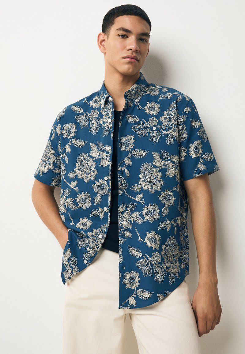 Camicia a maniche corte in tessuto blu scuro con motivi floreali beige, caratterizzata da una chiusura frontale con bottoni e orlo arrotondato, abbinata a pantaloni color crema.