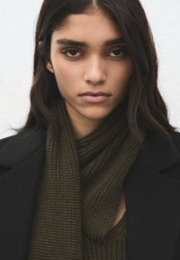 Ciemnozielony dzianinowy szalik wykonany z teksturowanej tkaniny, noszony z czarnym płaszczem, charakteryzujący się swobodnym układem i starannymi krawędziami.