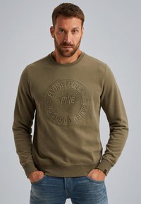 Olijfgroene sweatshirt met een gestructureerd circulair logo op de borst, ronde hals en geribbelde manchetten. Draagt met blauwe jeans.