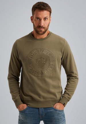Olijfgroene sweatshirt met een gestructureerd circulair logo op de borst, ronde hals en geribbelde manchetten. Draagt met blauwe jeans.