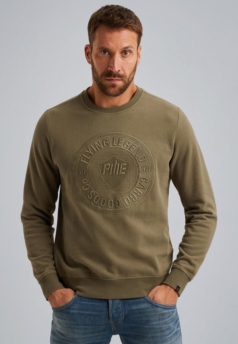 Olijfgroene sweatshirt met een gestructureerd circulair logo op de borst, ronde hals en geribbelde manchetten. Draagt met blauwe jeans.