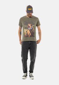 T-shirt en coton vert olive avec un imprimé graphique rétro et un jean fuselé gris foncé. Des baskets montantes noires complètent la tenue.