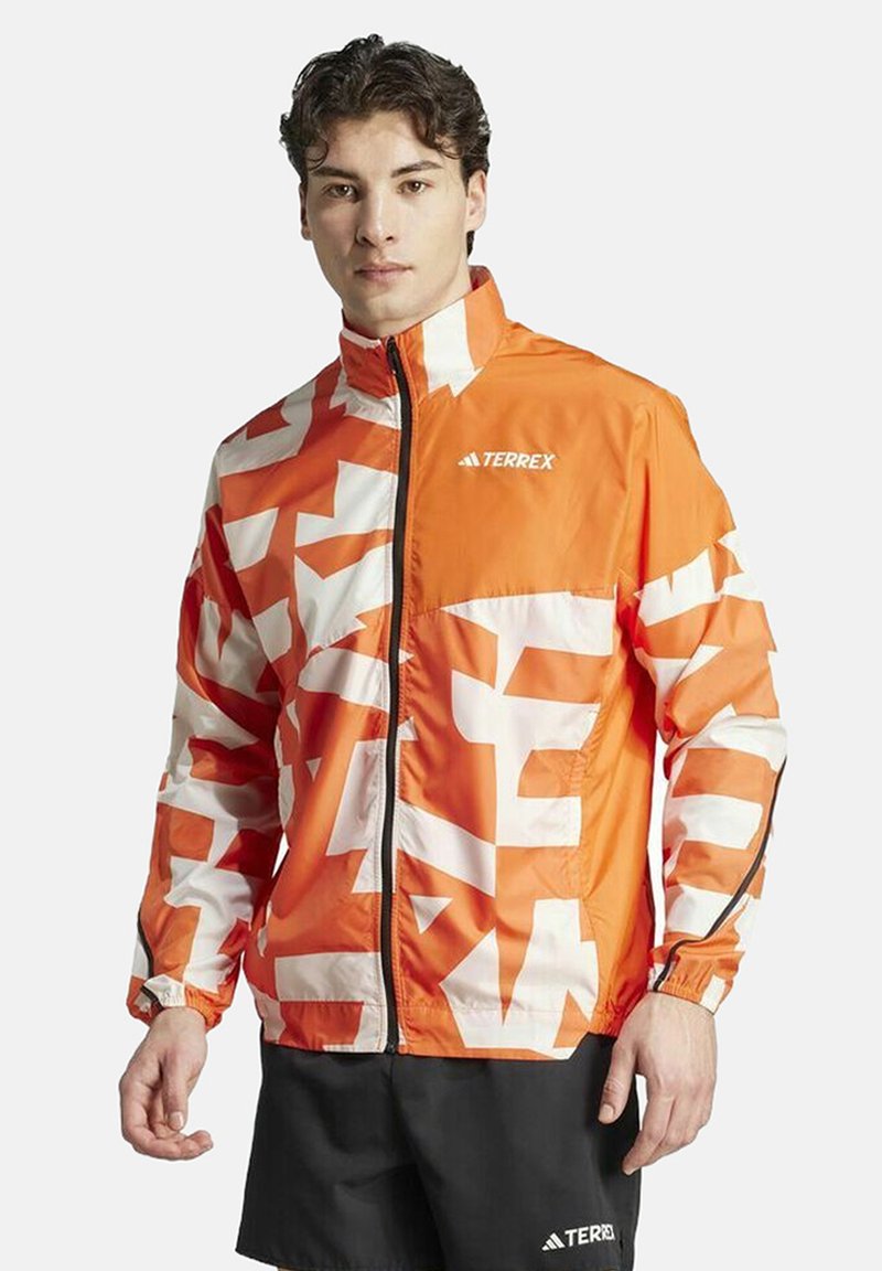Orange and white patterned Terrex jacket with a zip front, high collar, and elastic cuffs, paired with black shorts.