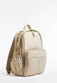 Beige ryggsäck tillverkad av vadderat tyg. Har justerbara remmar, en framficka med dragkedja, en sidoficka i nät och guldfärgade dragkedjor.