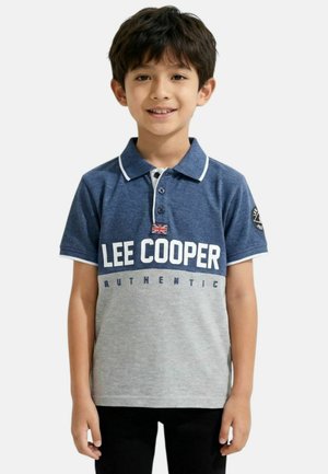 Garçon portant un polo Lee Cooper bleu et gris avec un drapeau Union Jack et un pantalon noir, debout et souriant sur un fond blanc.