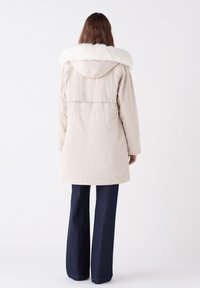Beige lange jas met een nepbont kraag, capuchon en elastische taille; gecombineerd met donkerblauwe flare jeans, met een gladde textuur en strakke lijnen.