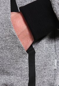 Hand in de zak van een grijs-zwart jasje met textuur, voorzien van een zwarte geribbelde boord en een klein Jack & Jones-label.