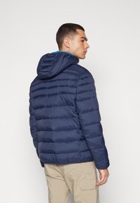 Vojska plava puffer jakna s kapuljačom, koja ima romboidni dizajn i glatku teksturu, nosi se preko bež hlača. Pogled s leđa naglašava kroj.