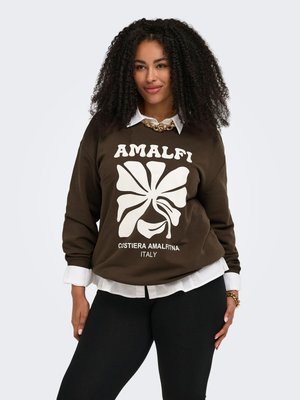 Kvinde med krøllet hår iført en brun "Amalfi Costiera Amalfitna Italy" sweatshirt over en hvid skjorte, sorte bukser og guldkæde.