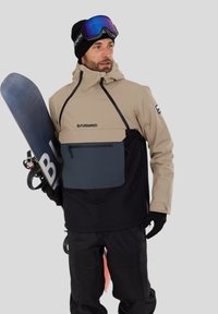 Snowboardová bunda v béžové a černé barvě s velkou přední kapsou. Texturovaný materiál se sportovním designem. Černé kalhoty a snowboard jsou součástí.