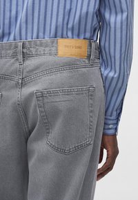 Personne portant un jean en denim gris avec une étiquette marron "Only & Sons" et une chemise manches longues rayée bleue, main détendue sur le côté.