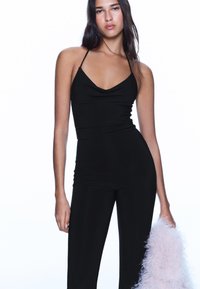 Svart ärmlös jumpsuit med draperad halsringning, figurnära silhuett och lätt rynkning vid midjan, kombinerad med en fluffig rosa accessoar.