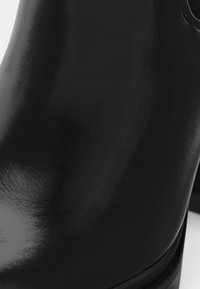 Primer plano de una bota de tobillo de cuero negro brillante con textura suave y detalles de punta redondeada.