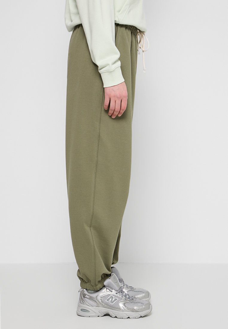 Personne portant un sweat-shirt vert clair, un pantalon de survêtement vert olive avec cordon de serrage et des baskets New Balance argentées, se tenant devant un fond blanc uni.
