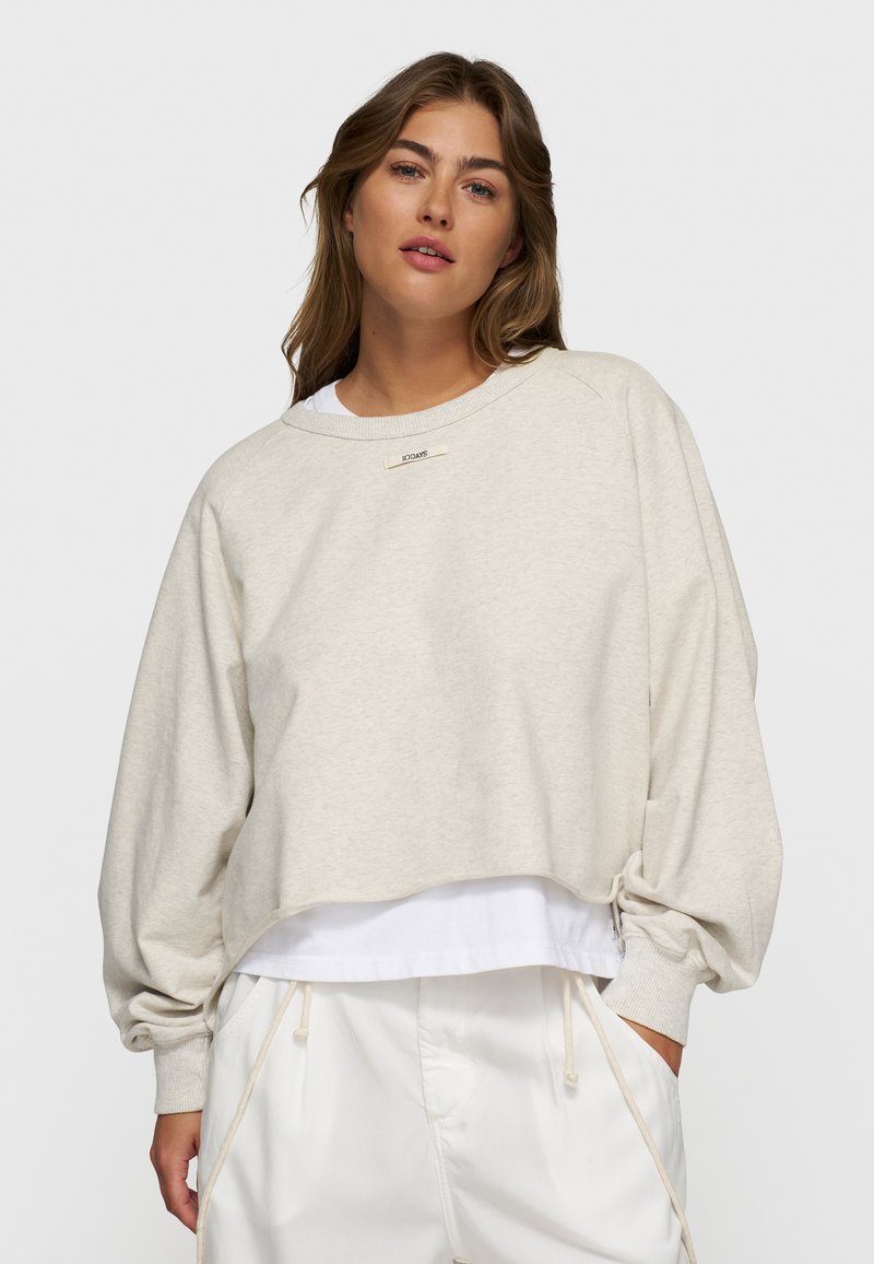 Felpa cropped beige chiaro con maniche a palloncino, caratterizzata da un collo a girocollo e un piccolo logo sul petto. Indossata sopra una camicia bianca.