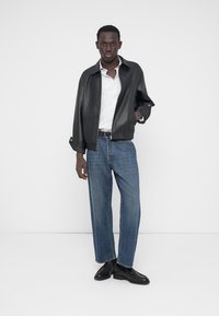 Veste en cuir noire portée sur une chemise blanche, associée à un jean droit bleu clair et des chaussures noires, sur un fond blanc uni.