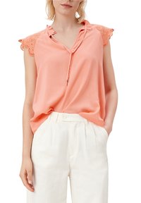 Blusa senza maniche color pesco con spalle rifinite in pizzo, scollatura a V e dettaglio con coulisse; abbinata a pantaloni bianchi larghi.
