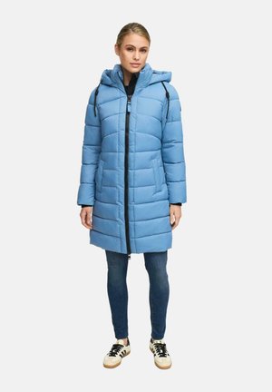 Vrouw in een lichtblauwe gewatteerde winterjas met capuchon, donkere jeans en beige sneakers met zwarte strepen, staand tegen een witte achtergrond.