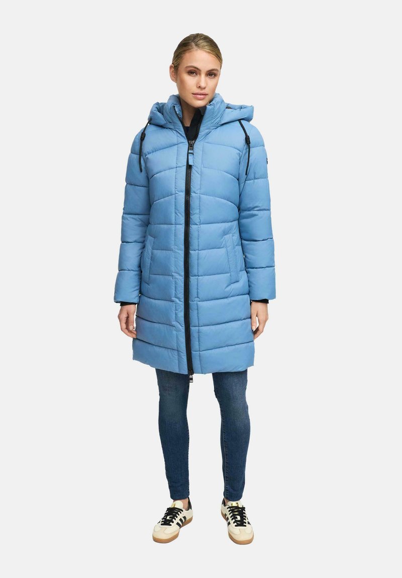 Vrouw in een lichtblauwe gewatteerde winterjas met capuchon, donkere jeans en beige sneakers met zwarte strepen, staand tegen een witte achtergrond.