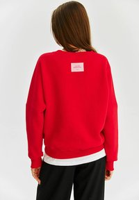 Červená veľká mikina s okrúhlym výstrihom, spadnutými ramenami, rebrovanými manžetami a lemom. Na zadnej strane sa nachádza štvorcová ružová značka.