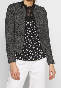 Sort-hvid jakke med fiskebensmønster og krave, lynlåsåben foran, båret over en sort bluse med blomsterprint, kombineret med hvide bukser.