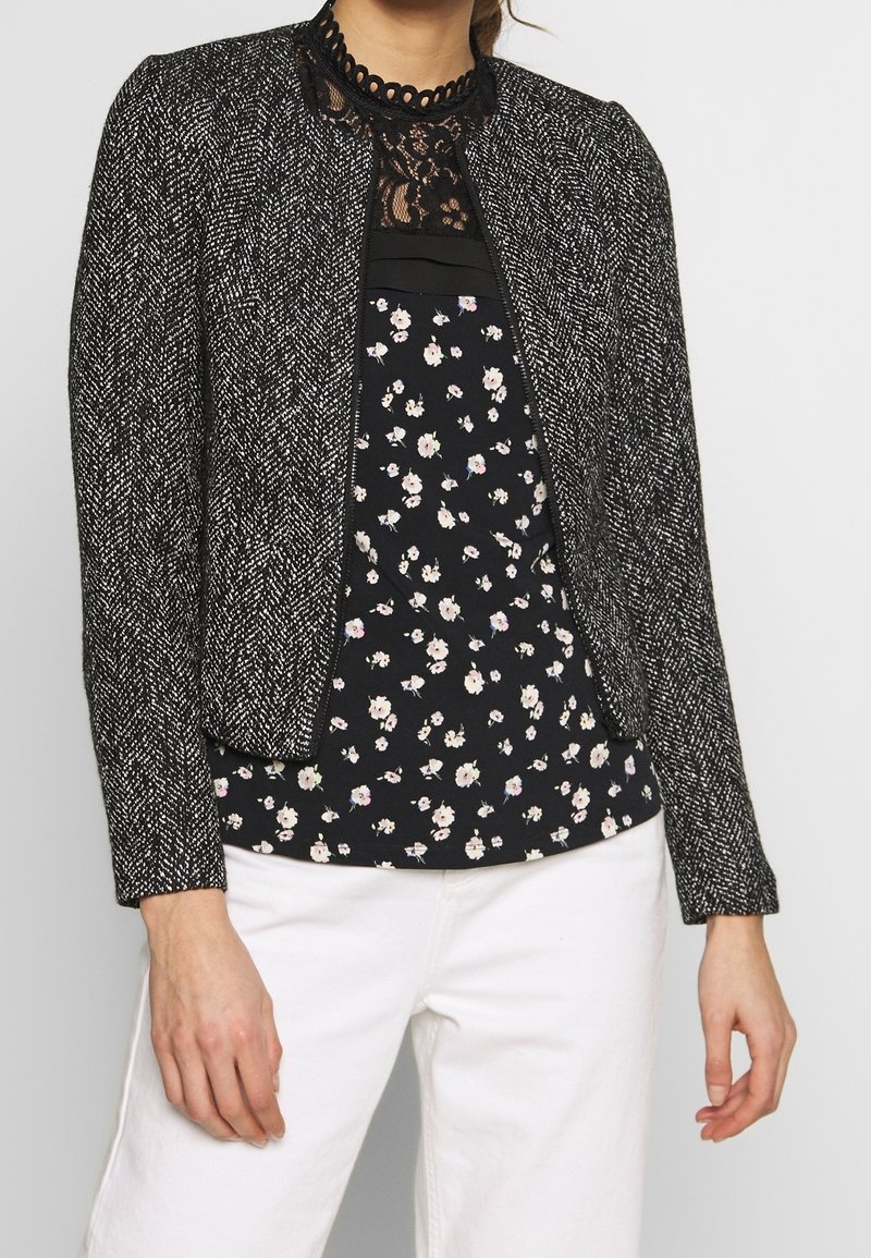 Sort-hvid jakke med fiskebensmønster og krave, lynlåsåben foran, båret over en sort bluse med blomsterprint, kombineret med hvide bukser.