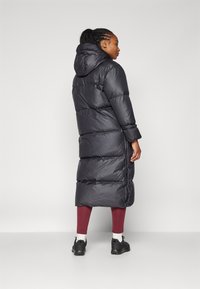 Lång svart puffjacka med huva, med horisontellt kviltade sektioner och sidofickor, tillsammans med vinröda leggings och svarta skor.