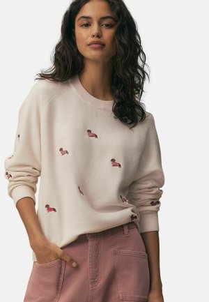 Woman wearing a cream sweater with embroidered dachshund dogs and mauve high-waisted pants with large front pockets, standing with one hand in pocket.