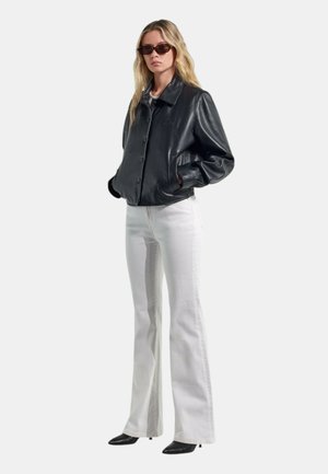 Donna con giacca di pelle scura, jeans bianchi a vita alta svasati, tacchi neri a punta e occhiali da sole, in piedi con le mani nelle tasche.