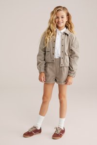 Tan krēpju tekstūras apgriezta jaka ar priekšējiem pogām, saskaņotiem šortiem un baltu krekliņu zemāk. Brūni sporta apavi un baltas zeķes noslēdz apģērbu.