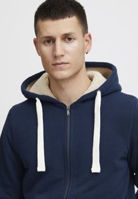 Granatowa bluza zapinana na zamek z beżowym kapturem wyłożonym polarem, białymi sznurkami do ściągania i gładką fakturą, prezentowana na jasnym tle.