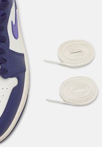 Baskets violettes et blanches avec dessus en cuir, accents en tissu texturé, et lacets plats violets à côté de deux lacets blancs enroulés avec embouts en plastique.