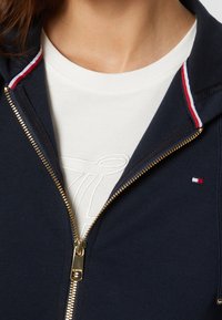 Sweat à capuche zippé bleu marine avec des détails rayés rouges et blancs au niveau du col, une fermeture éclair dorée, et un t-shirt crème avec un logo texturé en dessous.