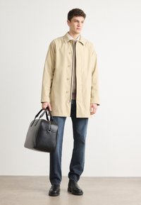 Μπεζ αδιάβροχο με γιακά και κουμπιά μπροστά, φορεμένο πάνω από ένα γκρι πουλόβερ. Σκούρα τζιν και μαύρες δερμάτινες μπότες, κρατώντας μια μαύρη τσάντα.