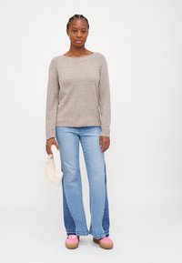 Maglione a maglia color marrone chiaro con scollatura rotonda, abbinato a jeans svasati blu. Borsa di peluche bianca tenuta nella mano sinistra, scarpe da ginnastica rosa ai piedi.