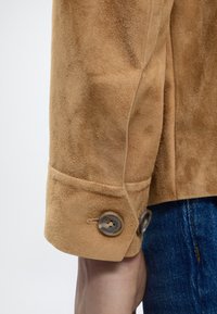 Κοντινό πλάνο του μανικιού ενός μπεζ σουέτ τζάκετ με δύο κουμπιά πάνω στον καρπό ενός ατόμου, σε συνδυασμό με μπλε τζιν.