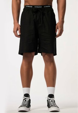Shorts noirs à cordon de serrage fabriqués en tissu léger, de longueur genoux, avec une taille élastique marquée, associés à des baskets montantes noires et des chaussettes blanches.