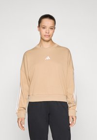 Beige hettegenser med rund hals og hvit Adidas-logo. Har hvite striper på lange ermer og en avslappet passform.