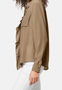 Blusa marrone a maniche lunghe con una texture liscia, caratterizzata da due tasche frontali e polsini con bottoni. L'orlo è leggermente curvato. Indossata con pantaloni bianchi.