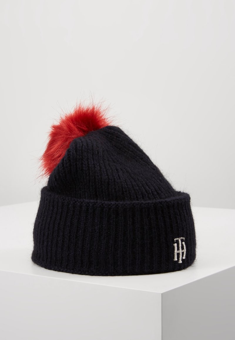 Bonnet en maille côtelée noire avec bord replié, pompon rouge duveteux sur le dessus et initiales blanches brodées sur le bord.