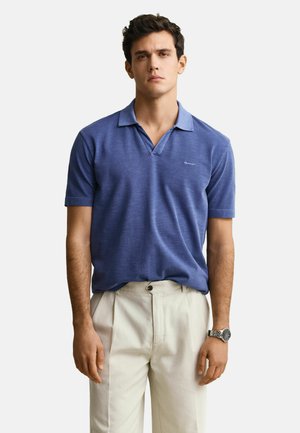Uomo che indossa una polo blu a maniche corte e pantaloni beige chiaro, in piedi con le braccia rilassate, indossa un orologio da polso al polso sinistro.