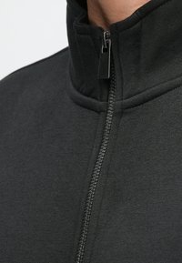 Lähikuva mustan takin vetoketjusta, joka on täysin suljettu korkealla olevassa kauluksessa, näkyvillä saumat ja teksturoitu kangas.