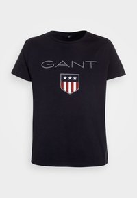 Svart bomullst-shirt med "GANT"-logotyp och ett skölddesign med tre stjärnor, samt röda och vita ränder i botten. Kort ärm, rund hals.