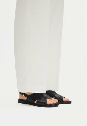 Pies con sandalias planas de cuero negro con tiras cruzadas, combinadas con pantalones blancos de pierna ancha, sobre un fondo blanco liso.