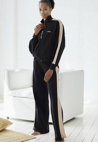 Schwarzer Sportanzug mit beigen Seitenstreifen, bestehend aus einer Reißverschlussjacke und passenden Hosen. Weicher Stoff und lockere Passform.