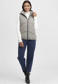 Sivi karirani prsluk s visokom ovratnikom, prednjim patentnim zatvaračem, kaki hlače i crne gležnjače, sa kviltanim uzorkom i bočnim džepovima.