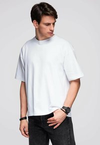 Młody mężczyzna w dużej, białej, gładkiej koszulce i czarnych dżinsach, spoglądający w dół w lewo, z jedną ręką w kieszeni.